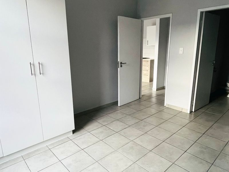A clean, empty bedroom featuring neutral tiled flooring and built-in cupboards, with direct access to an en-suite bathroom and adjacent living area.
