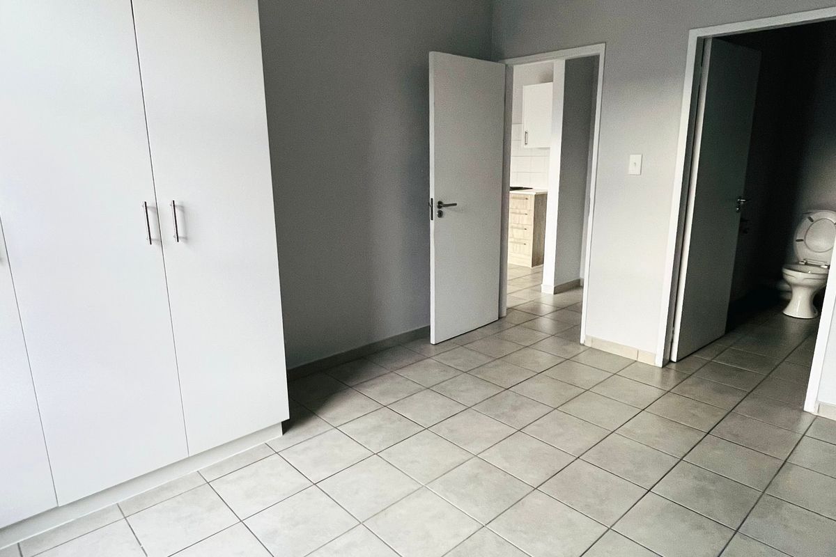 A clean, empty bedroom featuring neutral tiled flooring and built-in cupboards, with direct access to an en-suite bathroom and adjacent living area.