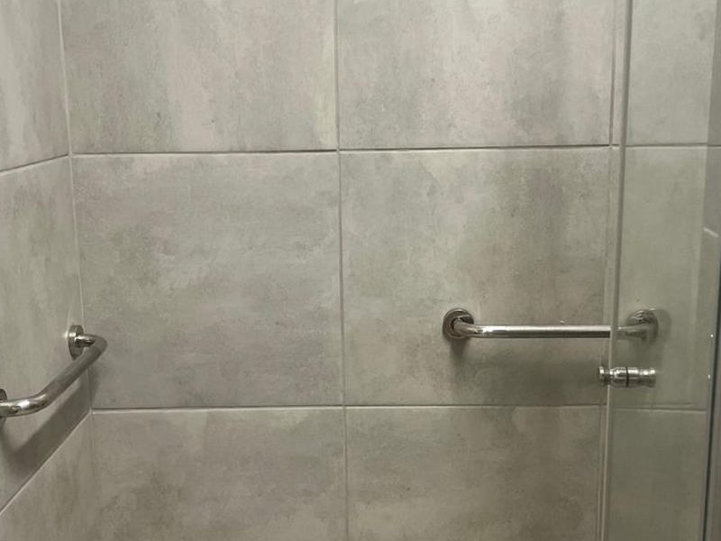 A modern shower enclosure featuring neutral ceramic wall tiles and installed stainless steel grab rails for accessibility.