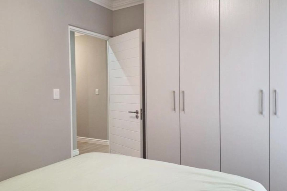 A clean, modern bedroom featuring neutral wall tones, built-in floor-to-ceiling cupboards, and a classic internal door leading to a hallway.
