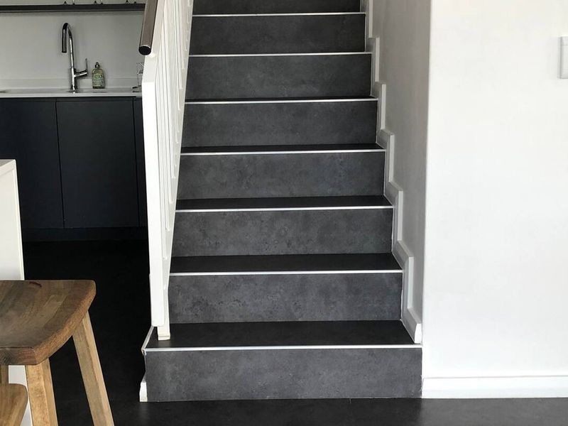 A modern interior view featuring a staircase with dark tiled steps and a white handrail, adjacent to a contemporary kitchen area.