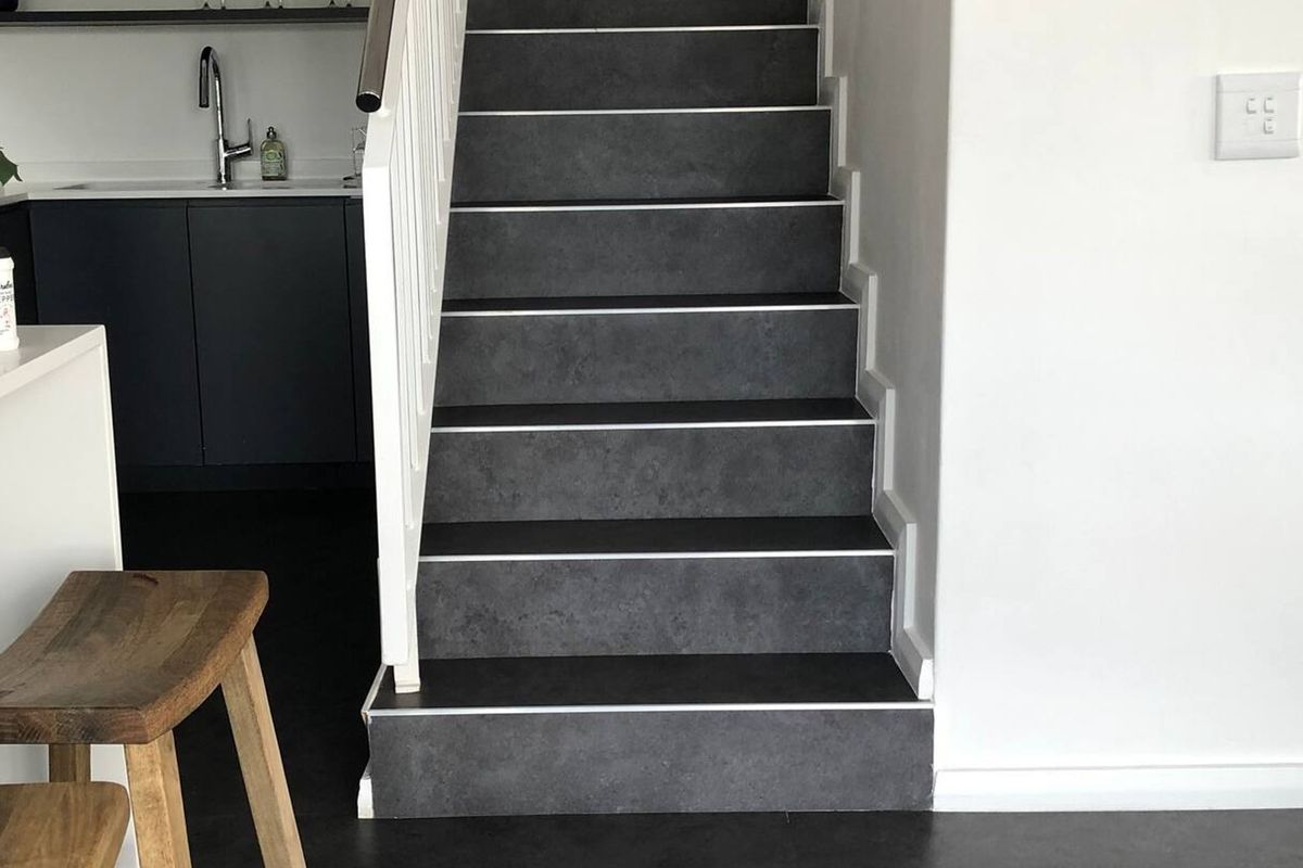 A modern interior view featuring a staircase with dark tiled steps and a white handrail, adjacent to a contemporary kitchen area.