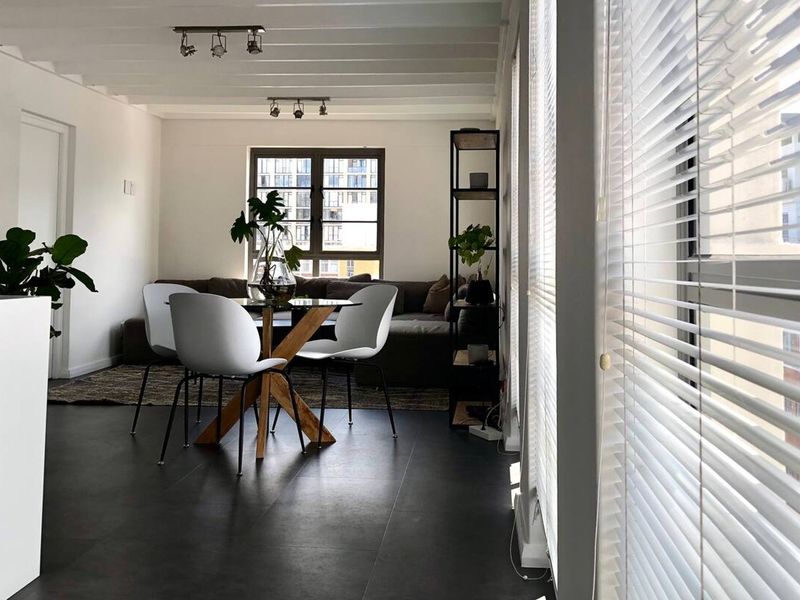 Bright open-plan living and dining area featuring modern tiled flooring, exposed beam ceilings, track lighting, and large windows with fitted venetian blinds.