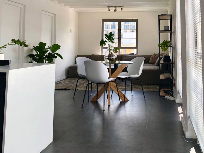 A bright open-plan living and dining area featuring modern dark floor tiles, neutral walls, and large windows with fitted venetian blinds.