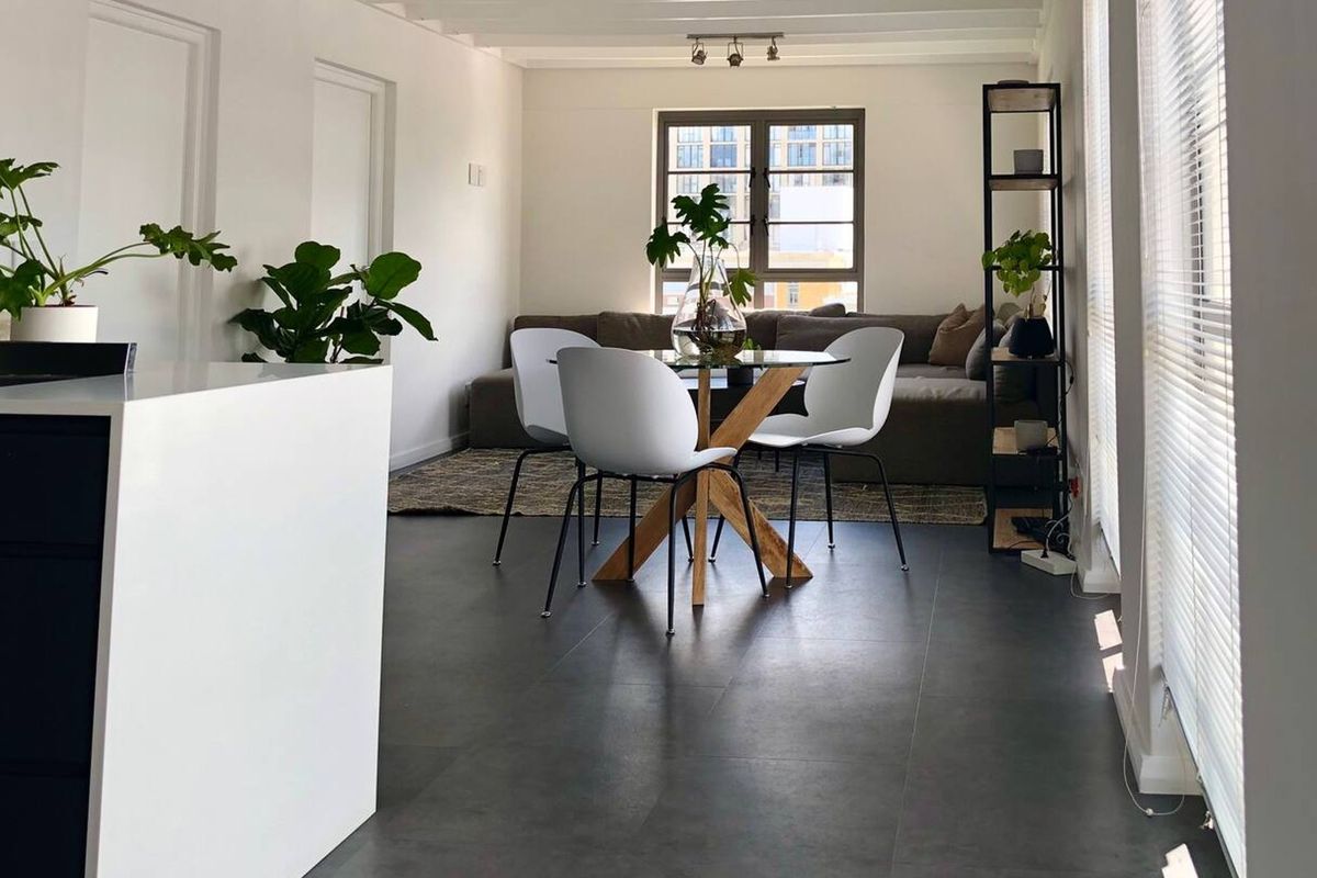A bright open-plan living and dining area featuring modern dark floor tiles, neutral walls, and large windows with fitted venetian blinds.