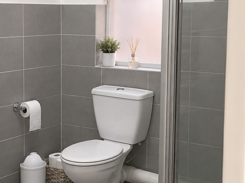 A modern bathroom featuring grey wall tiles, a white toilet, a washbasin, and a glass-enclosed shower area with a frosted window.