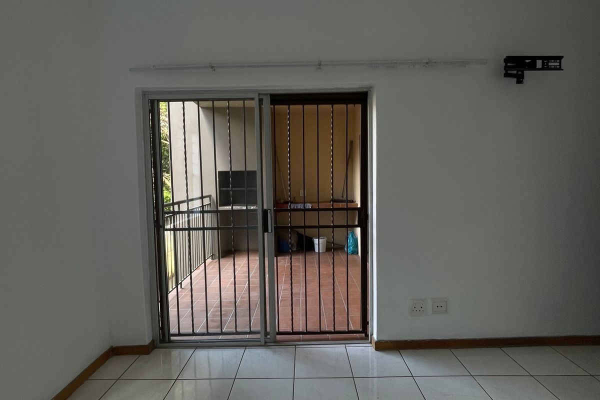 An empty living room featuring tiled flooring, a sliding glass door with security gate, and access to a balcony with a built-in braai.