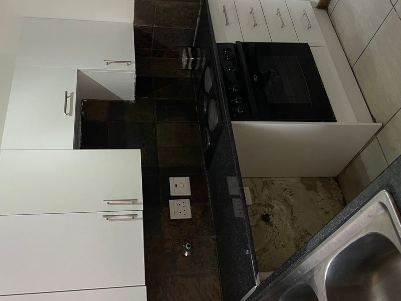 A compact kitchen featuring white cabinetry, dark stone countertops, a built-in oven, and a stainless steel sink, currently requiring minor finishing repairs.