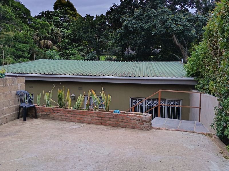 Rear exterior view of the cottage featuring a corrugated metal roof, concrete patio area, and brick retaining wall with metal security access.