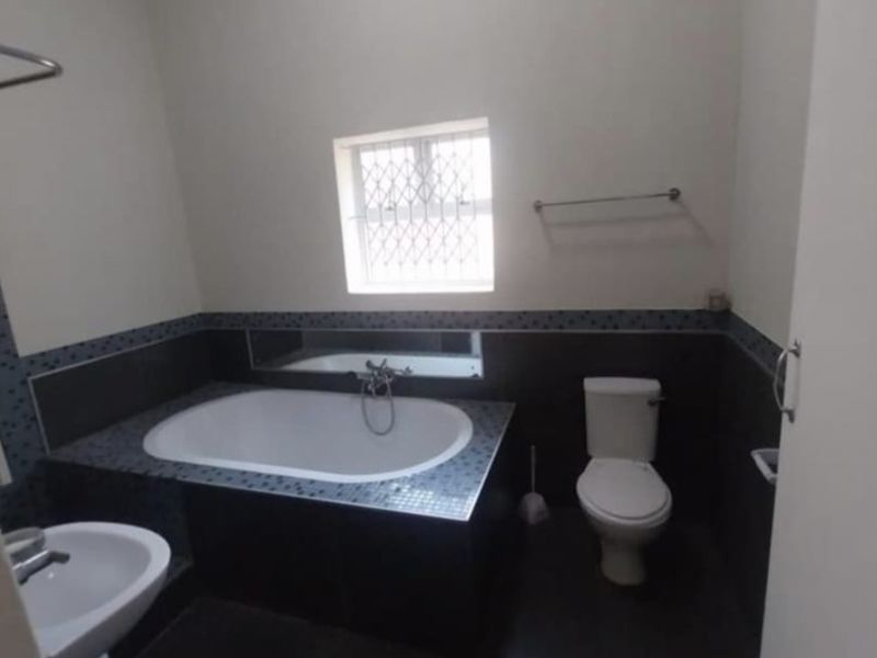 A functional bathroom featuring a built-in bathtub, pedestal basin, and toilet with dark mosaic tile accents and a window with security bars.