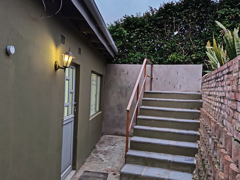 Rear exterior view of the cottage featuring a concrete staircase with metal handrail, brick retaining wall, and exterior wall lighting.