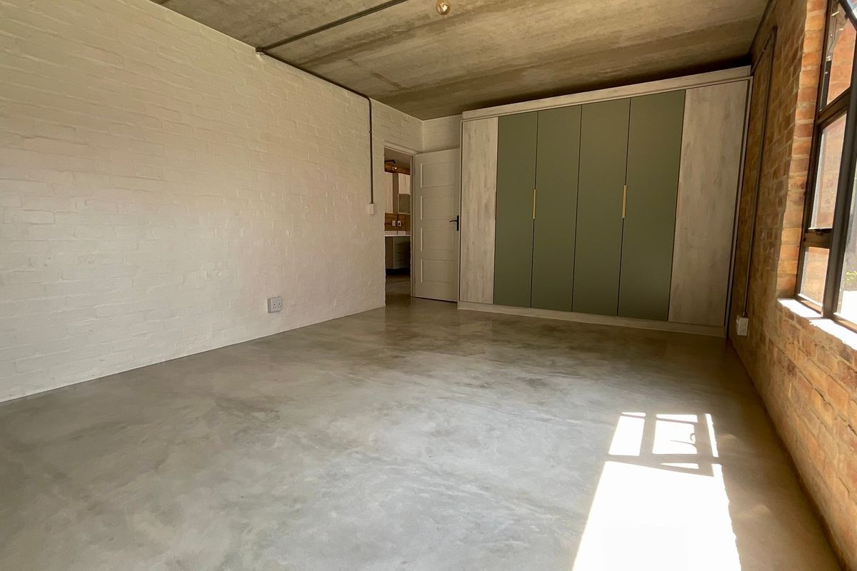 A modern bedroom featuring polished concrete floors, exposed brick and concrete elements, and a large built-in wardrobe with contemporary sage green cabinetry.