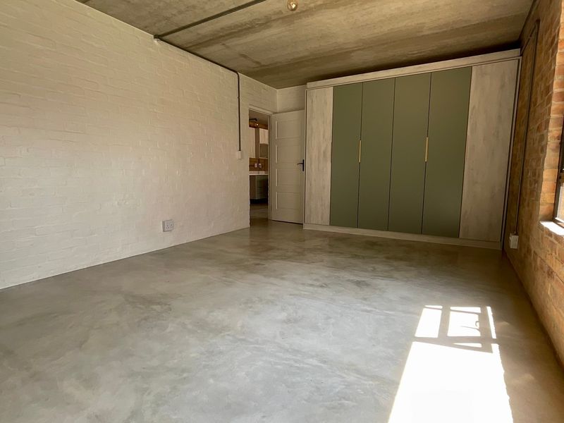 A modern bedroom featuring polished concrete floors, exposed brick and concrete elements, and a large built-in wardrobe with contemporary sage green cabinetry.