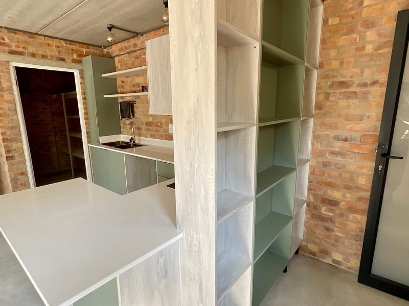 Modern industrial-style kitchen featuring exposed brick walls, polished concrete floors, sage green cabinetry, stone countertops, and integrated shelving.