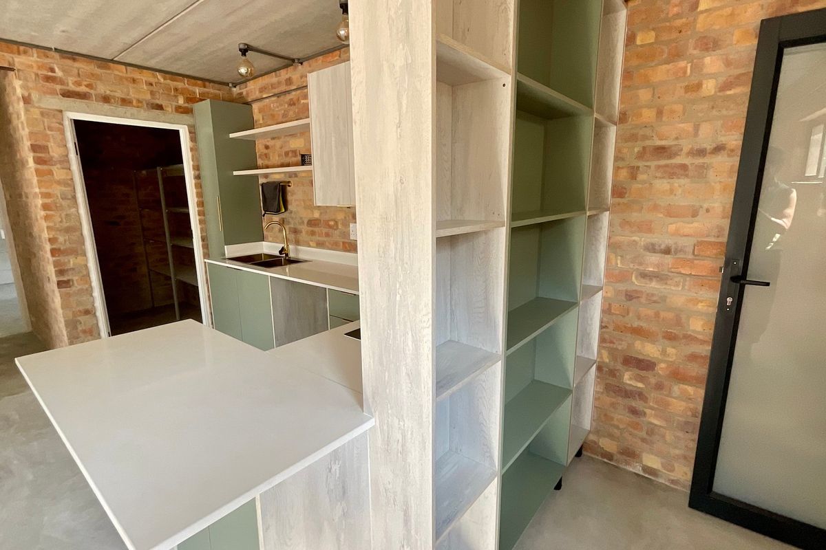 Modern industrial-style kitchen featuring exposed brick walls, polished concrete floors, sage green cabinetry, stone countertops, and integrated shelving.