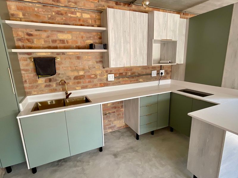 Modern industrial-style kitchen featuring exposed brick walls, sage green and wood-grain cabinetry, quartz countertops, and a sleek brass faucet.