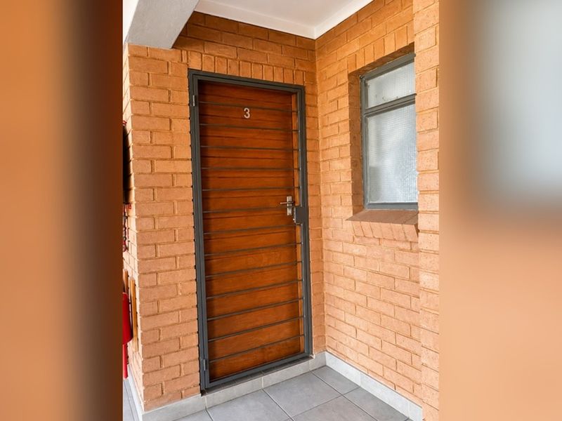 Entrance area of the apartment featuring a brick facade, tiled flooring, and a secure wooden door with a metal security gate.