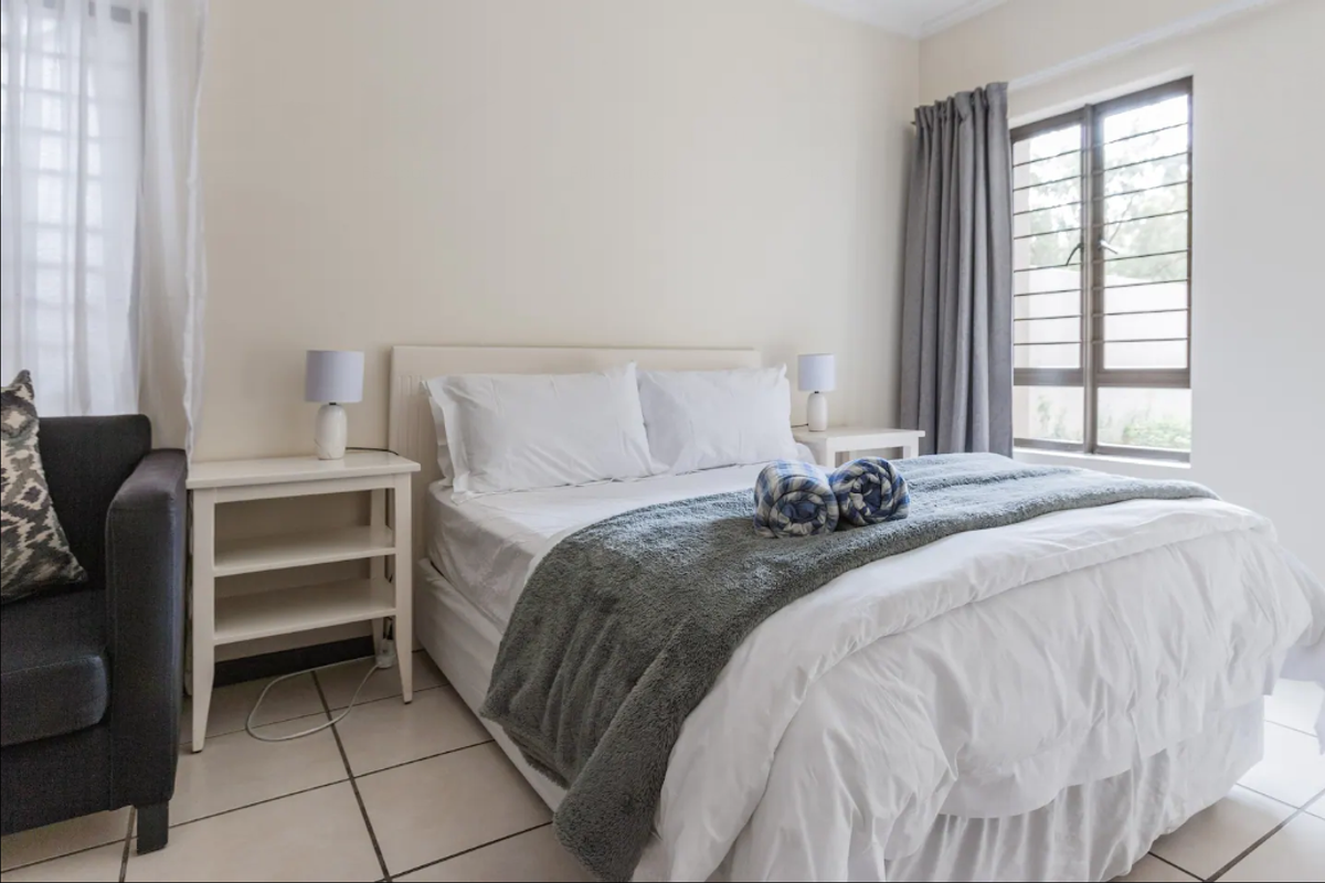 A well-lit bedroom featuring neutral wall tones, tiled flooring, and a window fitted with security bars.