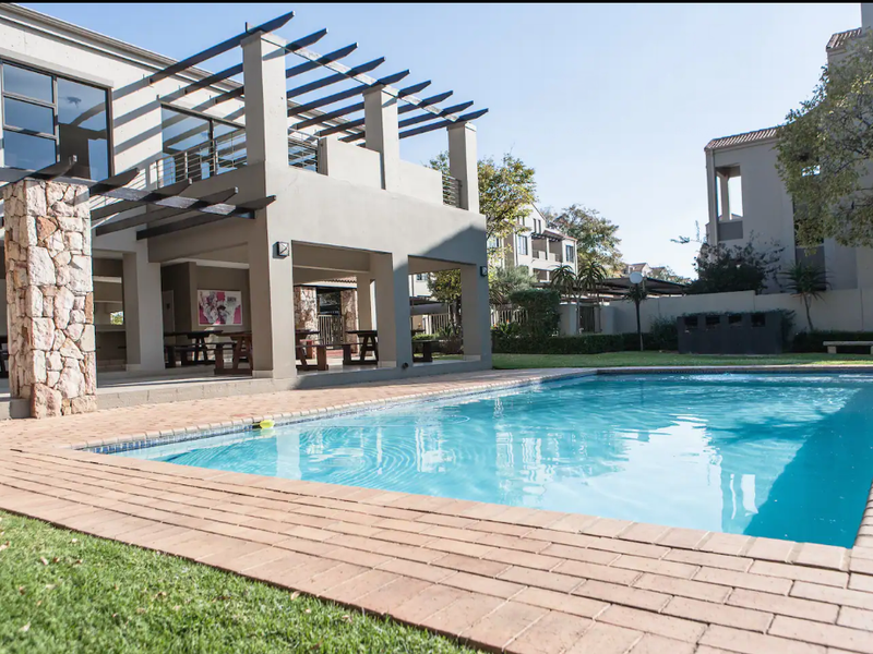A well-maintained communal pool area featuring brick paving, a covered patio with timber pergola, and stone-clad architectural details.