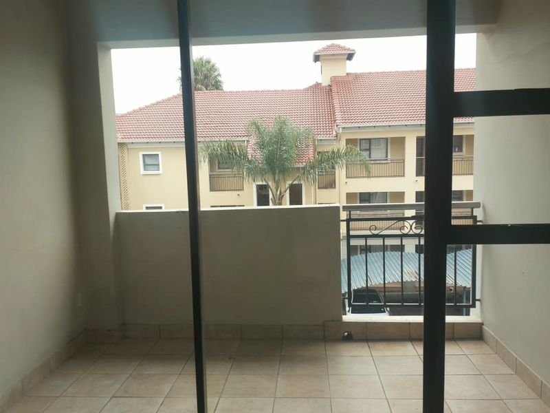 A covered balcony area featuring tiled flooring and a metal balustrade, offering an outlook over the apartment complex grounds.