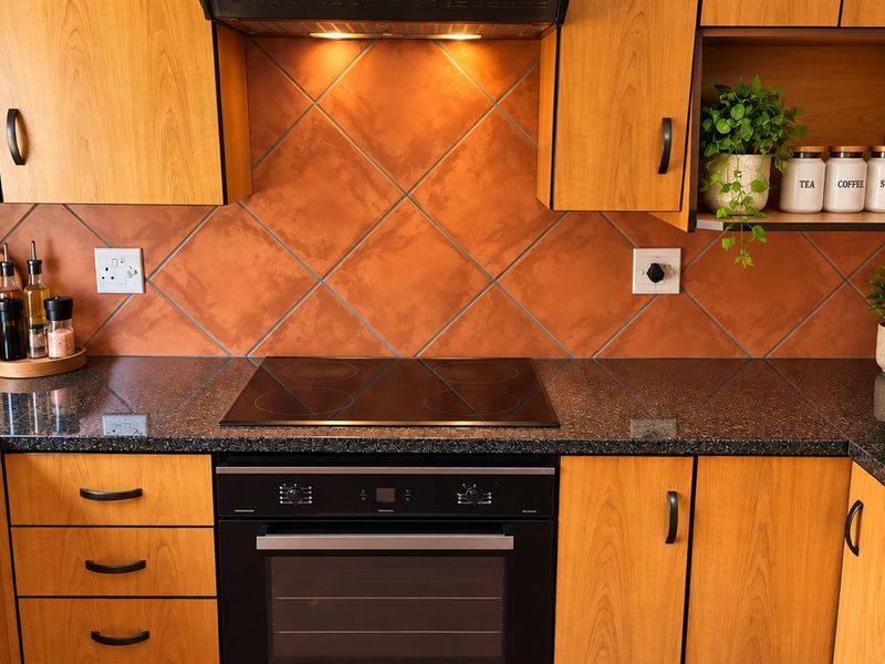 A functional kitchen featuring wood-finish cabinetry, dark granite countertops, a built-in electric oven and hob, and a decorative tiled backsplash.