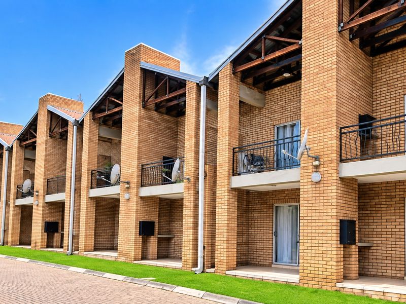 A row of modern face brick apartment units featuring exposed timber roof trusses, private balconies with metal railings, and paved driveway access.