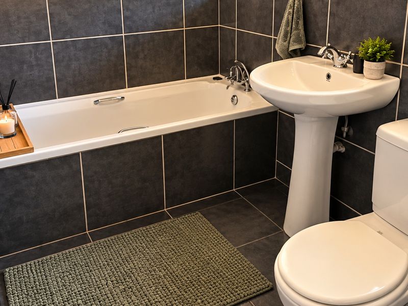 A modern bathroom featuring dark grey wall and floor tiles, a white bathtub, a pedestal basin, and a standard toilet.