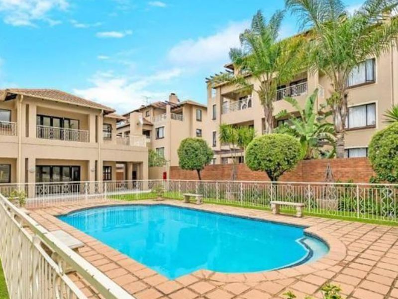 A well-maintained communal swimming pool area featuring paved surrounds, metal safety fencing, and landscaped gardens within an apartment complex.