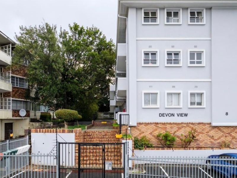 Front exterior view of Devon View apartment complex, featuring a multi-story building with brick and painted concrete facade, security fencing, and paved driveway access.