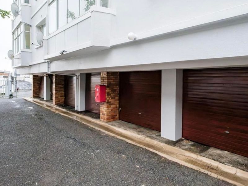 A row of secure, enclosed garage units located at the base of an apartment building with an asphalt driveway and fire safety equipment.