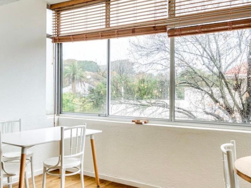 A bright living area featuring large windows with wooden blinds and light-toned laminate flooring, offering a pleasant view of the surrounding greenery.