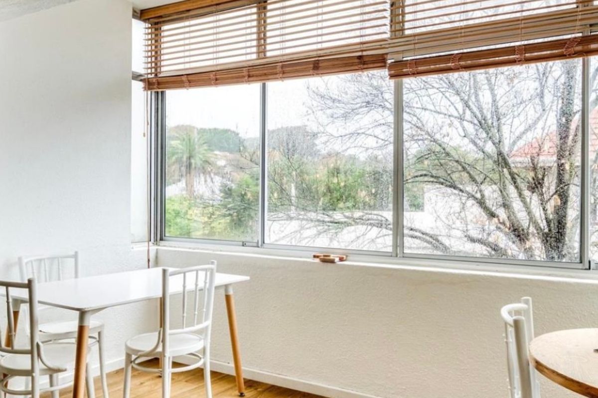 A bright living area featuring large windows with wooden blinds and light-toned laminate flooring, offering a pleasant view of the surrounding greenery.