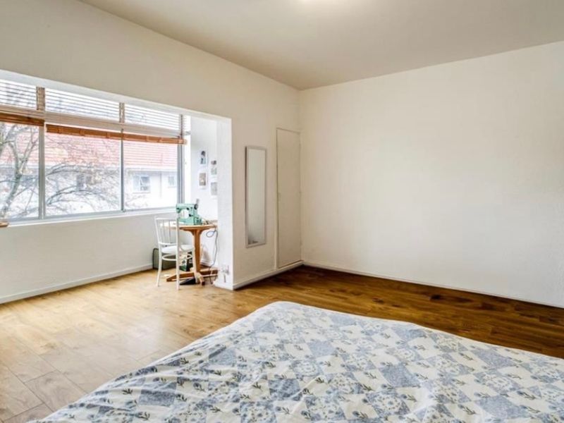 A bright and spacious bedroom featuring large windows, warm laminate flooring, and a built-in cupboard, offering a comfortable and well-lit living space.