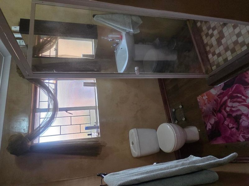 A functional bathroom featuring a shower enclosure, pedestal basin, and toilet, with security bars on the window and dated finishes throughout.