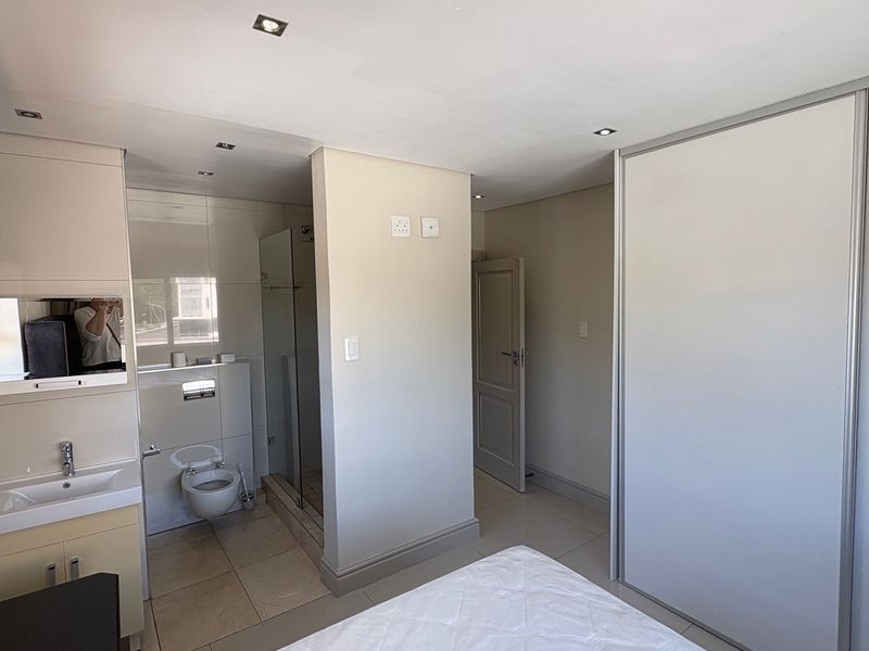 A modern bedroom featuring an open-plan en-suite bathroom with a glass shower, wall-mounted toilet, vanity basin, and large built-in sliding door wardrobes.