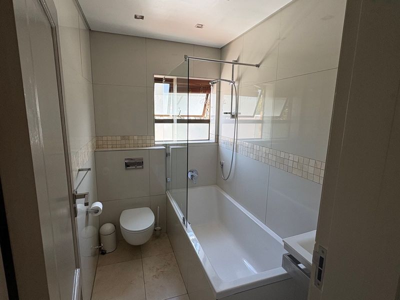 A modern, well-maintained bathroom featuring a wall-mounted toilet, a built-in bathtub with a glass shower screen, and neutral floor-to-ceiling tiling.