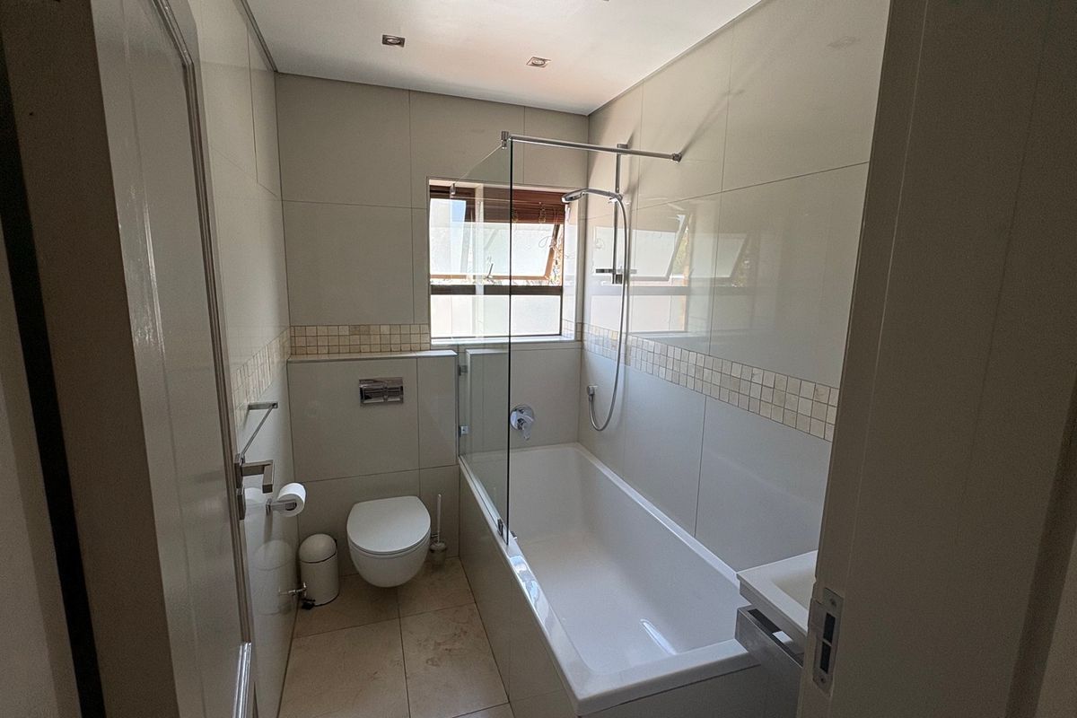 A modern, well-maintained bathroom featuring a wall-mounted toilet, a built-in bathtub with a glass shower screen, and neutral floor-to-ceiling tiling.