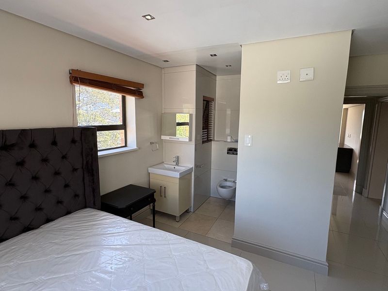 A bedroom featuring an open-plan en-suite bathroom with a vanity basin and wall-mounted toilet, complemented by tiled flooring and recessed lighting.