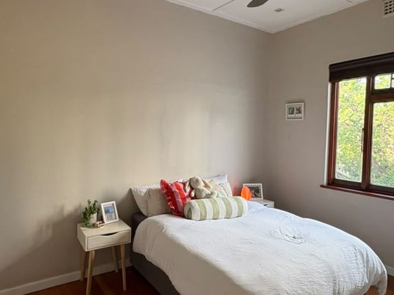 A well-lit bedroom featuring timber flooring, a ceiling fan, and a large window with a timber frame.