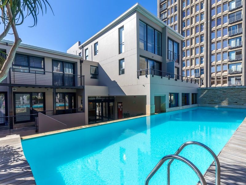 A well-maintained outdoor swimming pool area featuring timber decking, stone accents, and modern apartment building architecture with balconies.