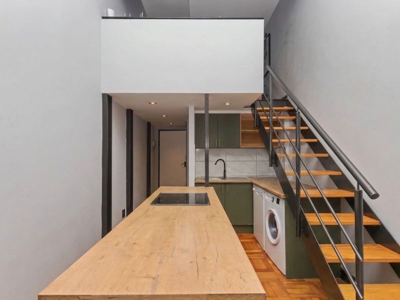 Modern open-plan kitchen area featuring olive green cabinetry, a wooden breakfast bar with integrated hob, and a staircase leading to a mezzanine level.