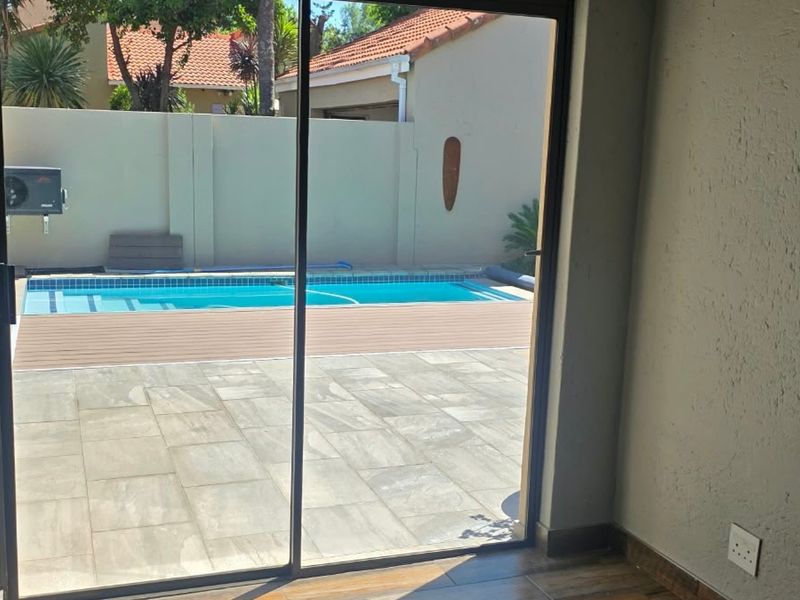 View from an interior space looking out through a sliding glass door onto a paved patio area with a swimming pool and boundary wall.