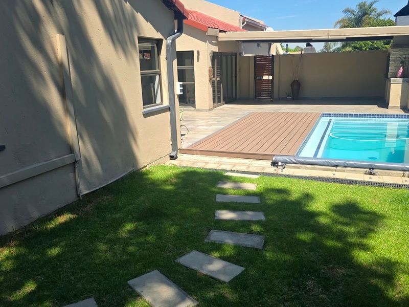 A well-maintained outdoor entertainment area featuring a swimming pool, wooden decking, a covered patio with a built-in braai, and a landscaped garden.