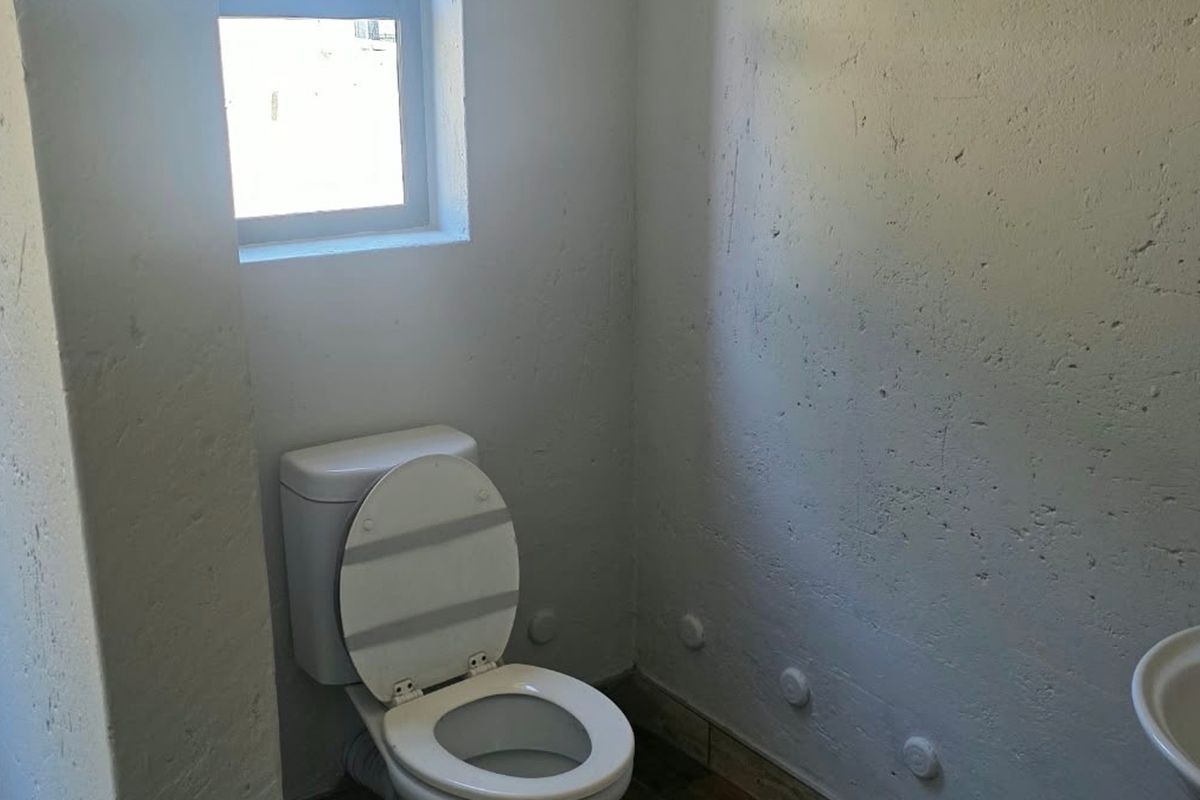 A basic, unfinished bathroom featuring a toilet, basin, and window, with rough wall textures and visible wall plugs requiring cosmetic attention.