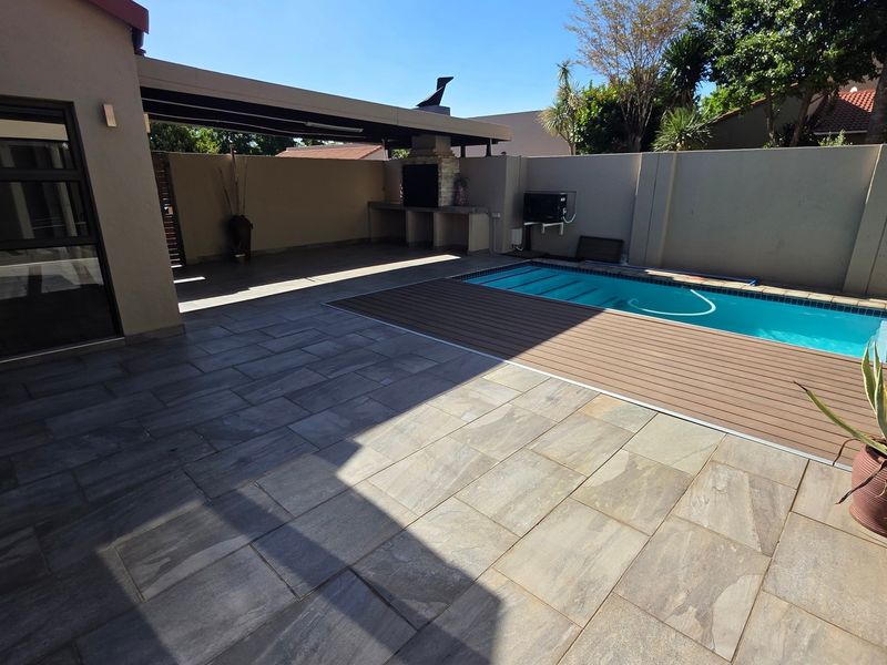 Outdoor entertainment area featuring a swimming pool, composite decking, stone patio, and a built-in braai under a covered patio structure.