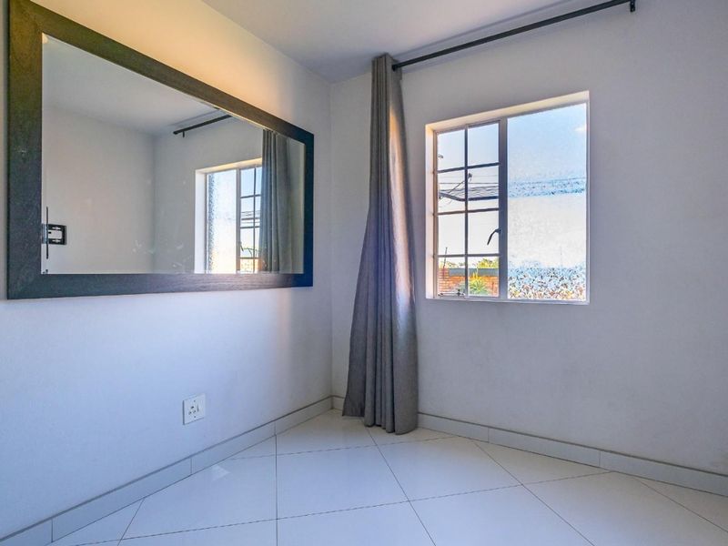A bright, empty bedroom featuring white tiled flooring, a window with curtain rod, and a large wall-mounted mirror.