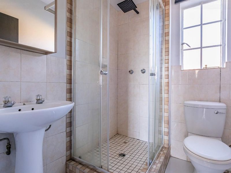 A functional bathroom featuring a pedestal basin, a glass-enclosed shower with a rain shower head, and a standard toilet with neutral wall tiling.