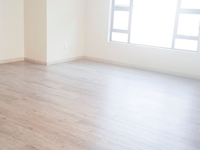 A bright, empty office space featuring light-toned laminate flooring, neutral walls, and a large window providing ample natural light.