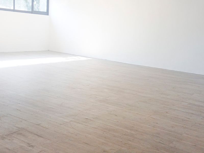 A bright, empty office space featuring light-toned laminate flooring and a large window, providing a clean and versatile environment for professional use.