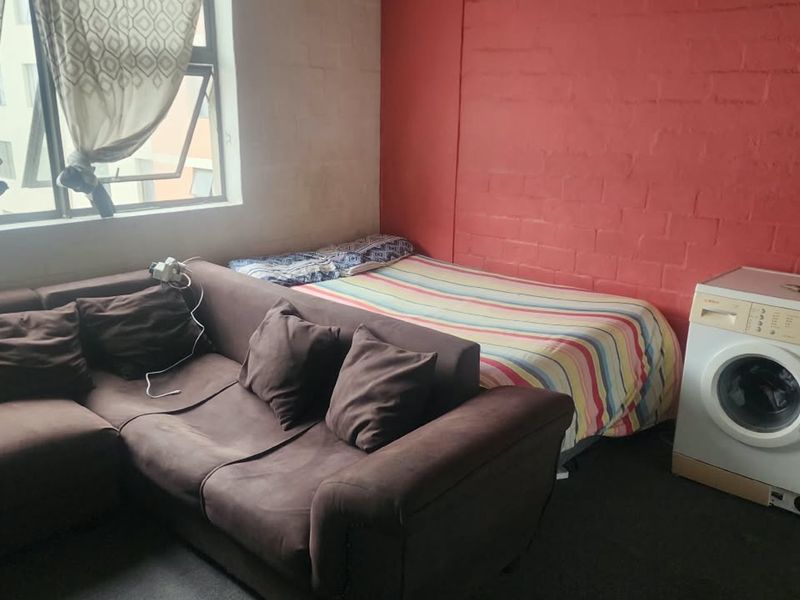 An open-plan living and sleeping area featuring an exposed red brick wall, a large window, and a dedicated space for laundry appliances.